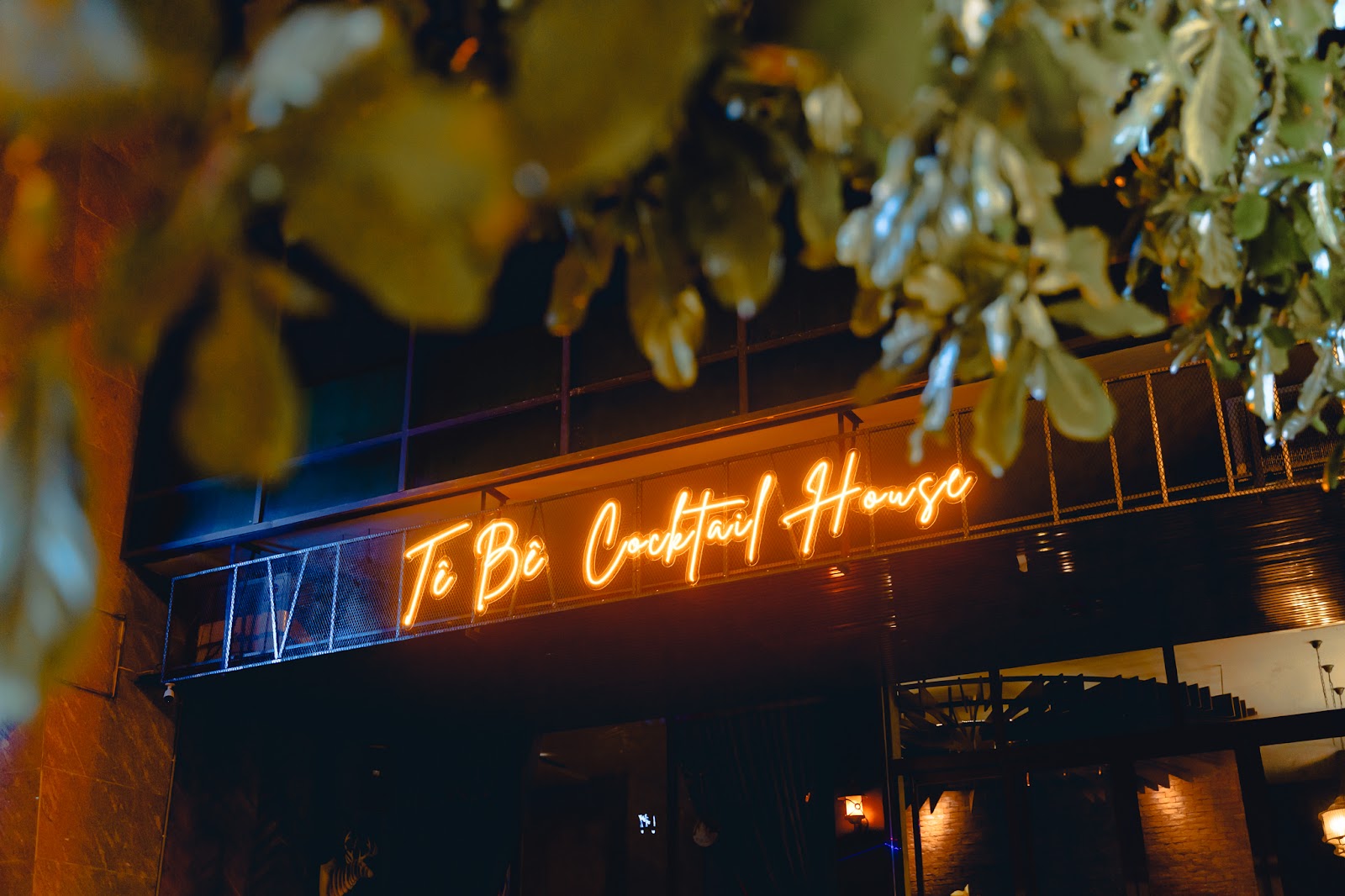 Biển đèn neon Tê Bê Cocktail House màu cam phía sau tán lá